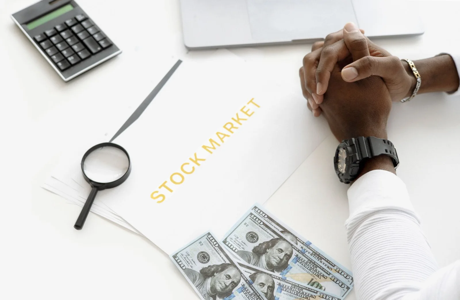 Mains croisées sur un dossier marqué stock market, à côté d'une loupe, d'une calculatrice et de billets